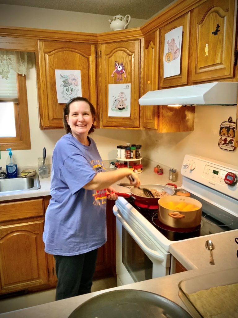 Terri, a white woman with brown hair smiles at the camera. She is in her kitchen cooking on the stove.